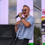 Mężczyzna w okularach śpiewa na scenie, ubrany w jasnoniebieską koszulę. Tło to duże ekrany LED z grafiką. Dzień, pochmurne niebo.