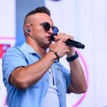 Mężczyzna w okularach i słuchawkach trzyma mikrofon. Ma na sobie jasnoniebieską koszulę. Tło jest rozmyte z elementami różu i bieli.