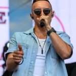 Na zdjęciu mężczyzna z mikrofonem śpiewa na scenie, ubrany w jasnoniebieską koszulę. Ma ciemne okulary i słuchawki douszne. Tło jest rozmyte, prawdopodobnie znajduje się na koncercie.