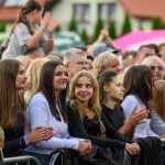 Grupa ludzi na imprezie na świeżym powietrzu, w jasnych i czarnych ubraniach. Tło to kolorowe namioty. Osoby wyglądają na zadowolone i klaszczą.