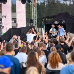 Na zdjęciu koncert na świeżym powietrzu, ludzie pod sceną podnoszą ręce. Na scenie muzycy z gitarą, z tyłu las. Dominuje kolor czarny i niebieski.