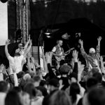 Na scenie muzycy grają koncert, otoczeni tłumem z uniesionymi rękoma. Tło jest ciemne, z widoczną konstrukcją sceny. Fotografia jest czarno-biała.
