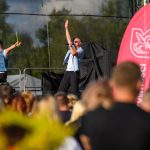 Na zdjęciu dwaj mężczyźni na scenie, jeden śpiewa. Obok nich kolorowe flagi, w tle zielone drzewa. Przed sceną widownia.