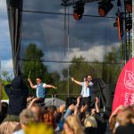 Dwóch mężczyzn w niebieskich koszulach występuje na scenie przed tłumem ludzi. Tło stanowi zielona przyroda i niebo. Po prawej stronie widoczne są czerwone i niebieskie flagi.