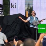Mężczyzna z gitarą w okularach śpiewa na scenie. Ma na sobie jeansową kamizelkę, z tyłu kolorowe światła. Przed nim tłum ludzi.