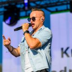 Mężczyzna w okularach i jasnej koszuli śpiewa na scenie, trzyma mikrofon. Jasne tło sceny z ekranem i reflektory.