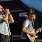 Dwóch mężczyzn na scenie. Jeden śpiewa do mikrofonu, drugi gra na błyszczącej gitarze. Tło jest ciemne, a ich ubrania jasne.