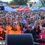 Tłum ludzi na wydarzeniu plenerowym w słonecznym świetle. Widoczne kolorowe namioty i atrakcje w tle. Atmosfera radosna, tłum jest zróżnicowany.