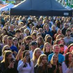 Tłum zgromadzony na zewnątrz. Ludzie robią zdjęcia i klaskają. W tle namioty i jasne kolory.