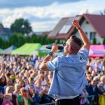 Mężczyzna śpiewa na scenie, trzyma mikrofon. Tłum ludzi, kolorowe namioty w tle. Jasny dzień, niebieskie niebo.