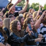 Grupa ludzi na koncercie, wznoszą ręce i robią zdjęcia. W tle zielone drzewa. Wiele osób ubranych w ciemne kolory.