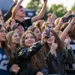 Grupa młodych ludzi z telefonami na koncercie, w radosnym nastroju. Kolorowe ubrania, oprawa w tle z zielenią. Tłum unosi ręce.