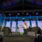 Trio mężczyzn występuje na scenie na świeżym powietrzu. Artyści noszą stroje w odcieniach niebieskiego i białego. Tło to kolorowe oświetlenie sceniczne.