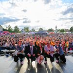 Na zdjęciu widoczna jest grupa muzyków na scenie i tłum ludzi na tle nieba z chmurami. Dominują kolory niebieskie, zielone i jasne. W tle widać atrakcje festynowe i namioty.