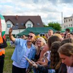 Mężczyzna robi selfie z grupą ludzi na festynie. W tle namioty i budynki, pochmurne niebo. Uczestnicy są uśmiechnięci i radośni.