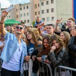 Mężczyzna w okularach robi selfie z grupą młodych ludzi za barierką. Uśmiechnięte twarze, różnorodne stroje, kolorowe tło. W tle budynki i namioty.