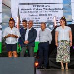 Grupa ludzi na scenie ubrana w casualowe i tradycyjne stroje. Tło to jasny ekran z tekstem. Pogoda jest pochmurna.
