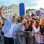 Młody mężczyzna w okularach przeciwsłonecznych robi selfie z grupą nastolatków. W tle kolorowe budynki i dmuchawce. Tłum ludzi uśmiecha się i pozuje.