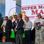 Grupa osób na scenie podczas ceremonii wręczenia nagród. Tło to ekran z logo SUPER MARKET MAJA. Osoby ubrani są elegancko, otoczeni są kolorowym oświetleniem.