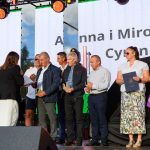 Grupa ludzi stoi na scenie, odbierając nagrody. Tło to duży ekran LED z jasnym niebem. Na zdjęciu dominują kolory: biały, czarny i fioletowy.