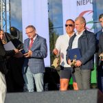 Na scenie grupa osób odbiera nagrody od kobiety w ciemnym stroju. W tle widoczny jest duży ekran i sprzęt oświetleniowy. Dominuje jasne oświetlenie i oficjalny charakter wydarzenia.