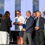 Grupa osób stoi na scenie, trzymając trofea. Tło to duży ekran, który wyświetla tekst. Atmosfera jest formalna, ale pogodna.