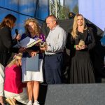 Grupa ludzi stoi na scenie na zewnątrz, w słońcu. Jedna kobieta wręcza mikrofon i nagrody. W tle niebieskie niebo i drzewa.