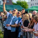 Na zdjęciu grupa osób robi selfie na tle zielonych namiotów. Ludzie stoją za barierkami, widać także budynki w tle. Dominuje kolor zielony i różnorodność ubrań uczestników.