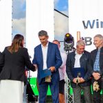Grupa ludzi stoi na scenie podczas ceremonii wręczenia nagród. Tło z jasnym niebem i białym banerem. Osoby trzymają niebieskie teczki i maskotki.