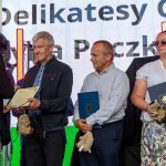 Na zdjęciu troje ludzi otrzymuje nagrody na scenie. Mają ozdobne dyplomy i pluszowe maskotki w rękach. Tło to jasny ekran z tekstem.