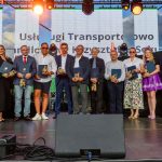 Grupa ludzi stoi na scenie z dyplomami i statuetkami, ubrana elegancko. Tło ozdobione kolorowymi światłami. Za nimi widoczne są trzy duże ekrany.