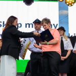 Na scenie kobieta przekazuje dokument innej kobiecie. W tle ludzie w mundurach na tle jasnego ekranu. Światła sceniczne są kolorowe.