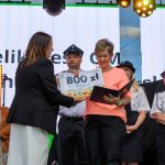 Na scenie kobieta w pomarańczowej bluzce odbiera nagrodę. W tle inne osoby w mundurach oraz ekran. Oświetlenie sceniczne podkreśla uroczystość.