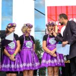 Grupa kobiet w fioletowych strojach i wiankach jest na scenie. Mężczyzna w garniturze trzyma mikrofon. Tło to biało-czerwony baner.