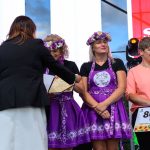 Dwie kobiety w fioletowych strojach z wiankami i jedna w pomarańczowej bluzce stoją na scenie. Jedna z nich trzyma tabliczkę z nagrodą. Tło jest biało-czerwone z napisem, na tle nieba.