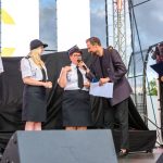 Na scenie widać cztery osoby, z których dwie noszą mundury. Mężczyzna trzyma mikrofon, a kobieta – teczkę. Tło stanowi czarne zadaszenie i plakat.