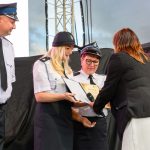 Na zdjęciu są umundurowani strażacy odbierający dyplom. Tło to scena z ciemnym zasłoną. Osoba w czerni wręcza nagrodę.