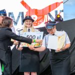 Na zdjęciu trzy osoby w mundurach strażackich odbierają nagrodę na scenie. Tło czarne z czerwono-białymi napisami. Uśmiechnięta kobieta trzyma czek na 500 zł.