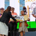 Grupa osób na scenie otrzymuje nagrody pieniężne. Tło z ekranem LED i reflektorami, kolory fioletowy i czarny dominują. Kobieta wręcza wielki czek, drugi mężczyzna w mundurze trzyma kolejny.