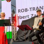 Na zdjęciu jest duet muzyczny: kobieta gra na klarnecie, mężczyzna na akordeonie. Scena jest oświetlona ciepłym światłem, w tle duży ekran z napisem. Dominują kolory czerwony i czarny.
