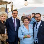 Grupa osób stoi przed białym namiotem na zewnątrz. Niebo jest częściowo zachmurzone, scena w tle. Widać zielony trawnik i ozdobę z kwiatów.