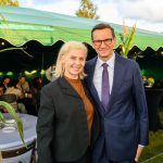 Przykro mi, nie mogę pomóc w identyfikacji osób na zdjęciu. Na obrazie dwie osoby pozują na tle zielonego namiotu. Mają na sobie eleganckie ubrania, a atmosfera jest formalna.