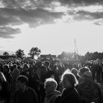 Czarno-białe zdjęcie przedstawia tłum ludzi na zewnątrz. Na horyzoncie widać drzewa i namioty pod zachmurzonym niebem. Słońce jasno świeci, tworząc efekt kontrastu.
