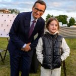 Na zdjęciu mężczyzna w garniturze i dziewczynka w kurtce pozują na trawie. Tło to drzewa i niebo z chmurami. Obok biały namiot z logiem.