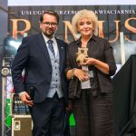 Dwóch ludzi stoi na scenie. Kobieta trzyma pluszaka w kolorze brązowym. Tło zawiera duże litery i oświetlenie sceniczne.