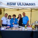 Grupa ludzi stoi pod białym namiotem z napisem KGW Ulijanki. Na stole są przetwory, soki i ciasta. Tło kremowe, dekoracje z wieńcami.