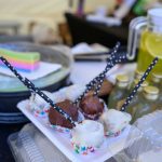 Kolorowe cake popsy z białą i czekoladową polewą na talerzu. W tle słoiki z żółtym napojem i naczynia. Jasne, rozmyte tło.