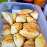 Plastikowe pudełko pełne złocistych empanad. W tle butelki napojów gazowanych. Sceneria kuchenna o żywych kolorach.