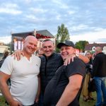 Trzech mężczyzn uśmiecha się na tle sceny plenerowej. Niebo jest zachmurzone, a trawa zielona. W tle widoczni ludzie i domy.
