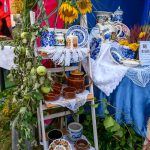 Stojak z ceramiką na tle niebiesko-różowego namiotu. Dekoracje z żółtymi kwiatami i zielonymi liśćmi. Klasyczne naczynia z wzorami.